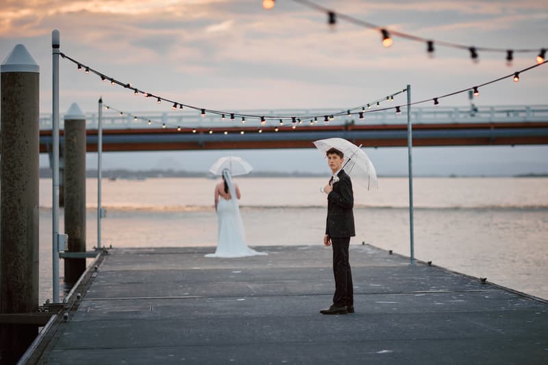 The groom stands on a pier holding a transparent umbrella and looking towards the camera, while the bride stands further away facing the water, also holding a transparent umbrella, at Sandstone Point Hotel.