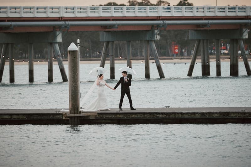 The bride Terion and groom Tyler hold hands and walk on a dock over water at Sandstone Point Hotel, each holding a transparent umbrella. A bridge is visible in the background.