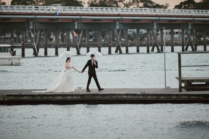 The bride Terion and groom Tyler walk hand in hand on a dock at Sandstone Point Hotel, each holding a transparent umbrella with water in the background and a bridge behind them.