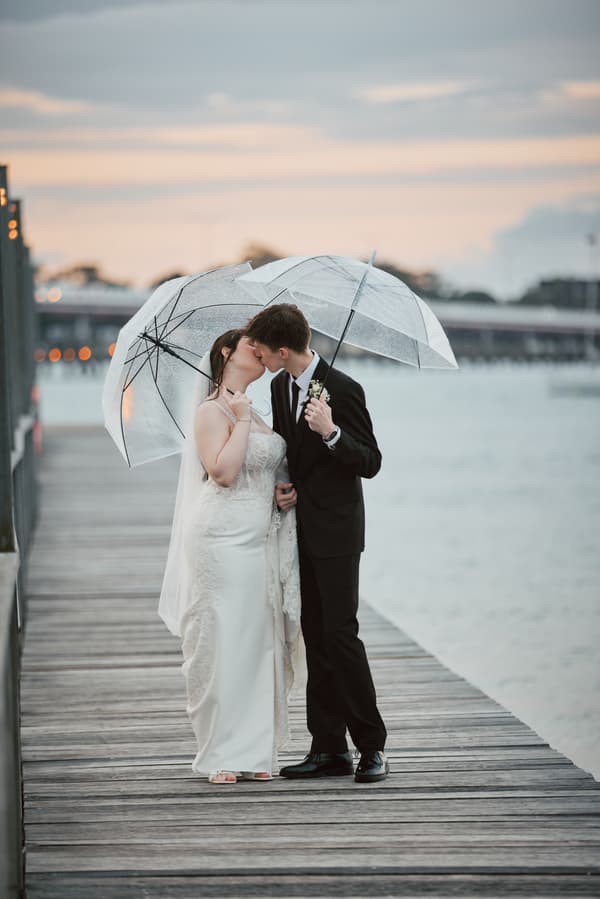 Terion the bride and Tyler the groom kiss under clear umbrellas on a wooden pier at Sandstone Point Hotel during their couple portraits session.