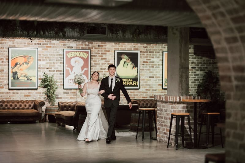 Terion the bride and Tyler the groom walk arm in arm through the reception area at Sandstone Point Hotel — The Cellar, with brick walls, vintage posters, and leather couches in the background.