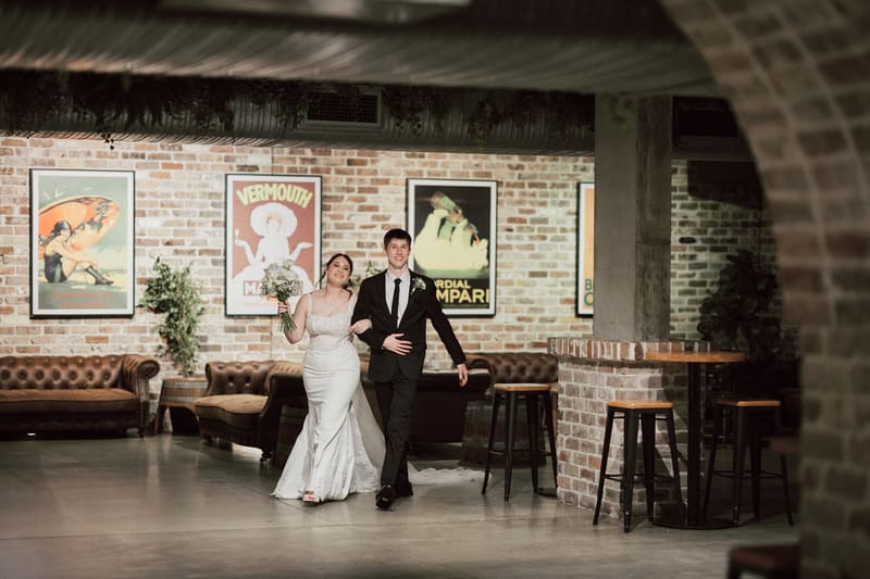 Terion the bride and Tyler the groom walk arm in arm through the reception area, with brick walls, vintage posters, and leather couches in the background. Wedding photography add-ons by She Said Yes.