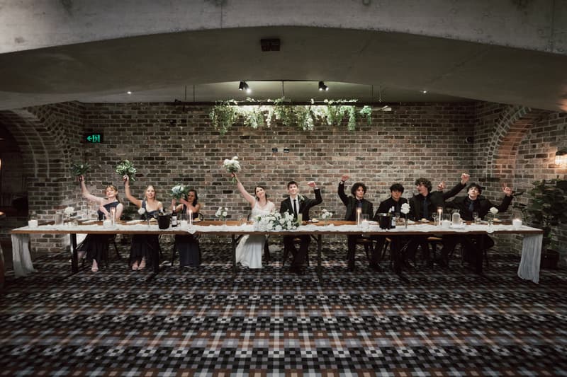 Terion the bride and Tyler the groom sit at the center of a long head table at Sandstone Point Hotel — The Cellar, flanked by bridesmaids and groomsmen raising their hands and bouquets in celebration.
