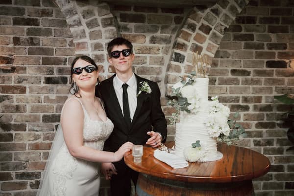 Bride Terion and groom Tyler wearing sunglasses stand beside a three-tier white wedding cake decorated with white flowers on a wooden barrel table at Sandstone Point Hotel — The Cellar.