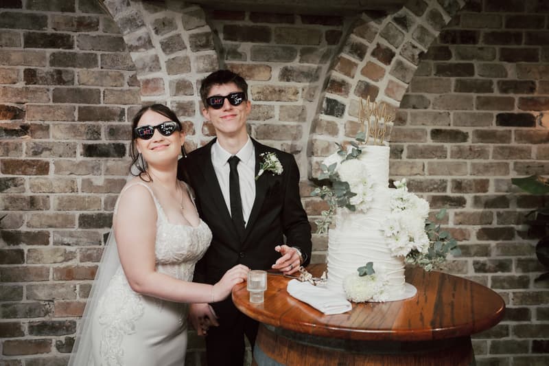 Bride Terion and groom Tyler wearing sunglasses stand beside a three-tier white wedding cake decorated with white flowers on a wooden barrel table at Sandstone Point Hotel — The Cellar.