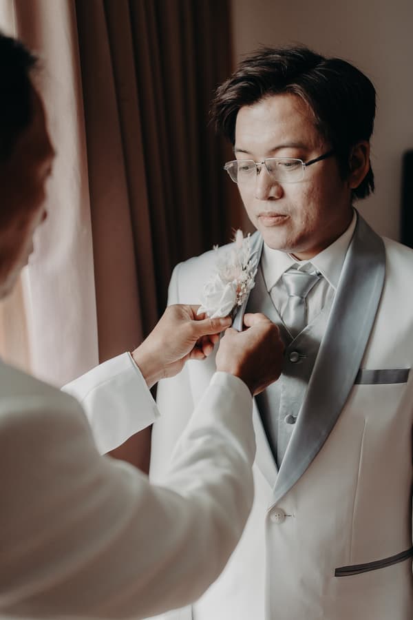 The groom Timothy stands while another man adjusts his boutonniere near a window with curtains at Royal on the Park.