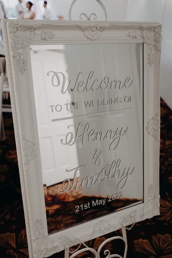 Decorative white-framed mirror sign welcoming guests to the wedding of Henny and Timothy dated 21st May 2022, placed on a stand with a patterned carpet floor visible.