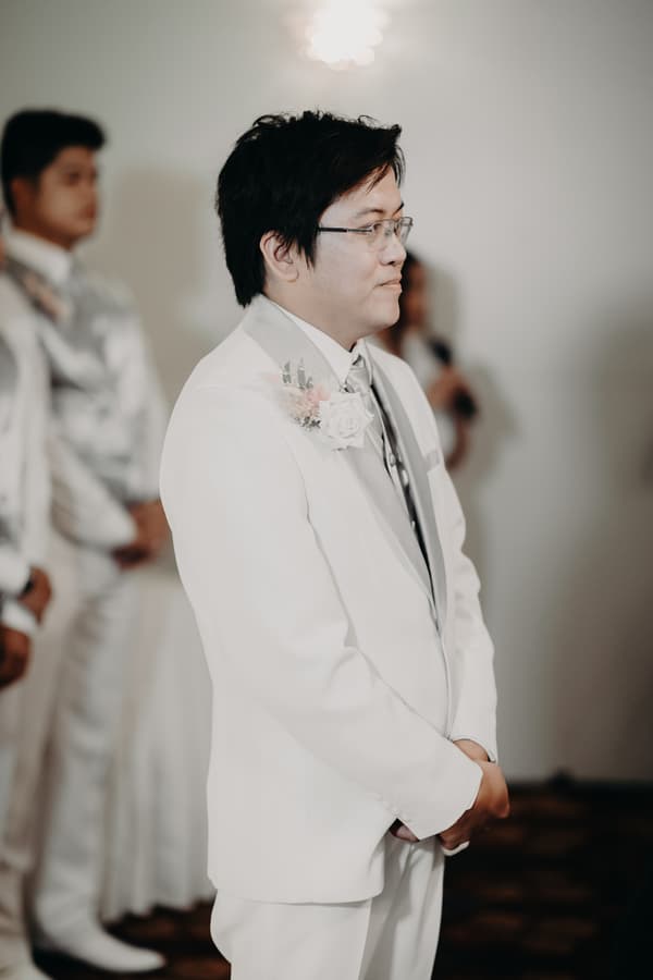 The groom Timothy stands in a white suit with a boutonniere at the ceremony stage at Royal on the Park, with groomsmen blurred in the background.