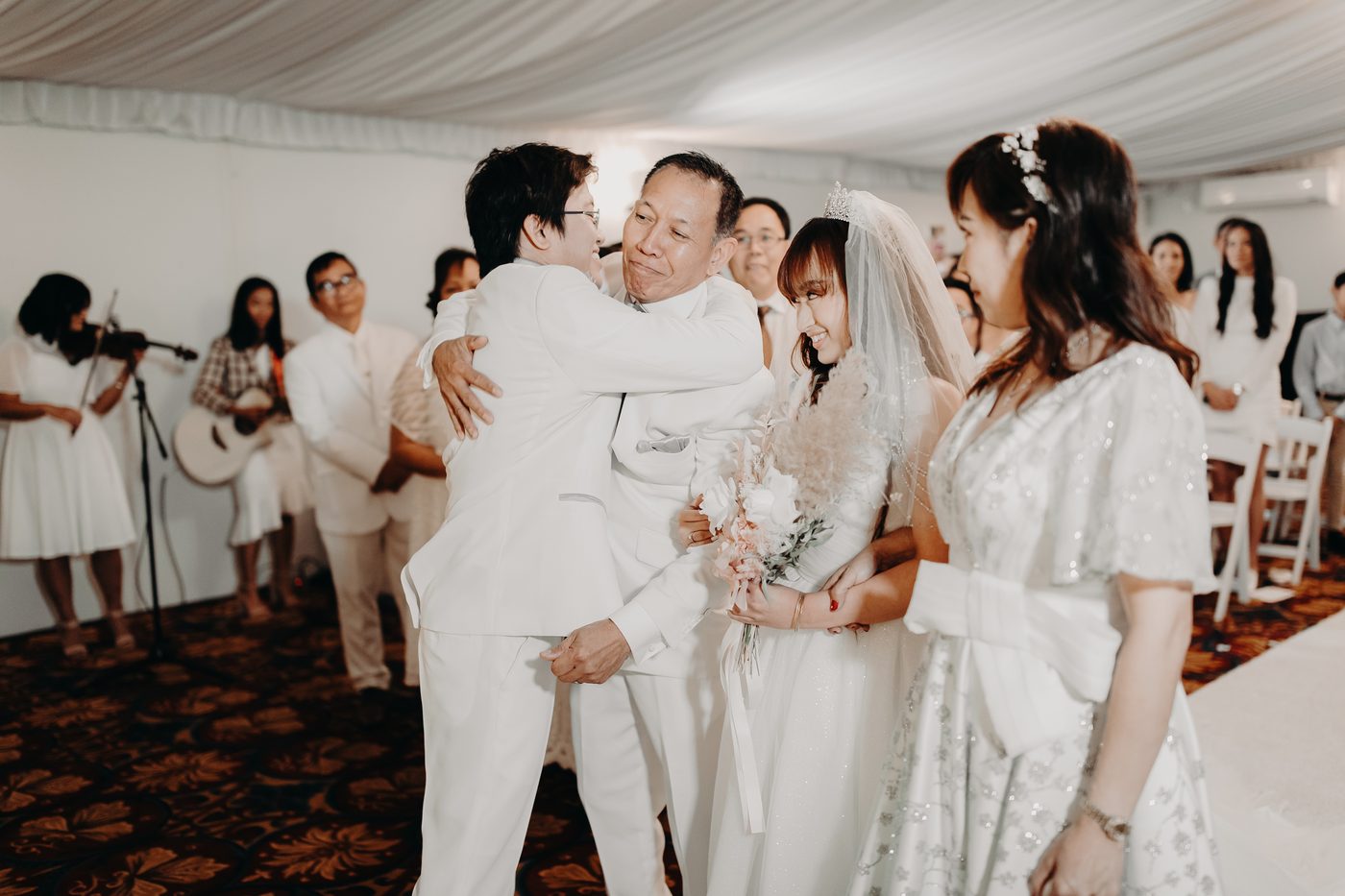 Timothy hugs an older man while Henny, the bride, stands beside them holding a bouquet at the ceremony stage. A woman in a silver dress stands nearby, and musicians play violin and guitar in the background. Wedding photography pricing estimate by She Said Yes.