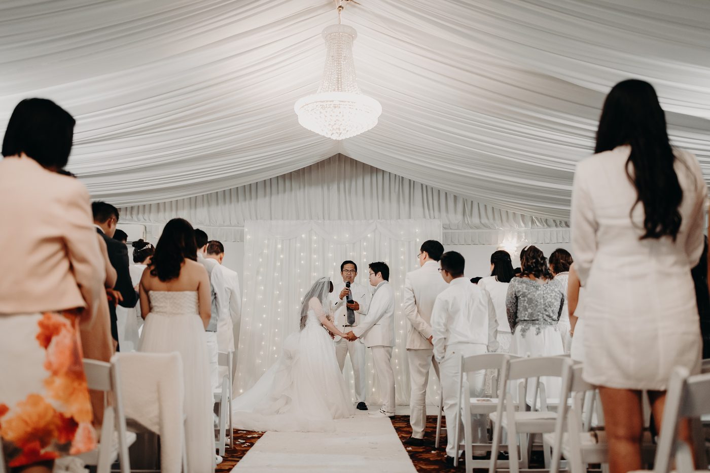 Bride Henny and groom Timothy hold hands facing the officiant during their wedding ceremony, surrounded by standing guests in a white draped venue. Elopement photographer Brisbane by She Said Yes.