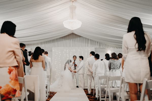 Bride Henny and groom Timothy hold hands facing the officiant during their wedding ceremony at Royal on the Park, surrounded by standing guests in a white draped venue.