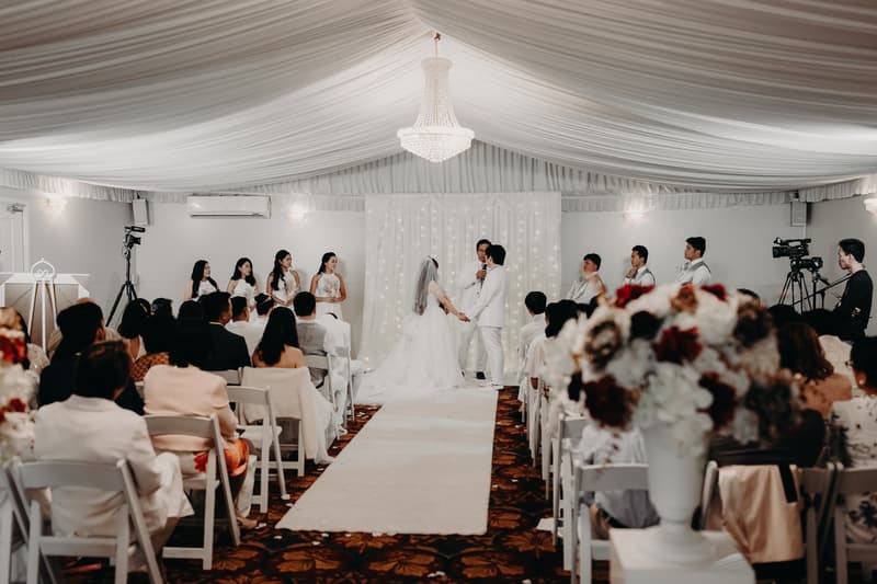 Bride Henny and groom Timothy hold hands facing the officiant during their wedding ceremony at Royal on the Park, with bridesmaids and groomsmen seated and standing on either side and guests watching from rows of chairs.