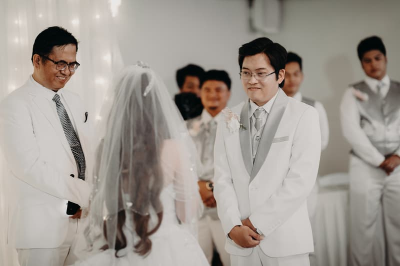 Timothy and Henny stand facing each other at the ceremony stage at Royal on the Park, with groomsmen in silver vests and white shirts standing behind Timothy.