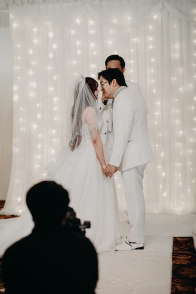 Bride Henny and groom Timothy hold hands and lean in for a kiss during their wedding ceremony, with the officiant standing behind them and a photographer capturing the moment. Gold Coast wedding videographer by She Said Yes.