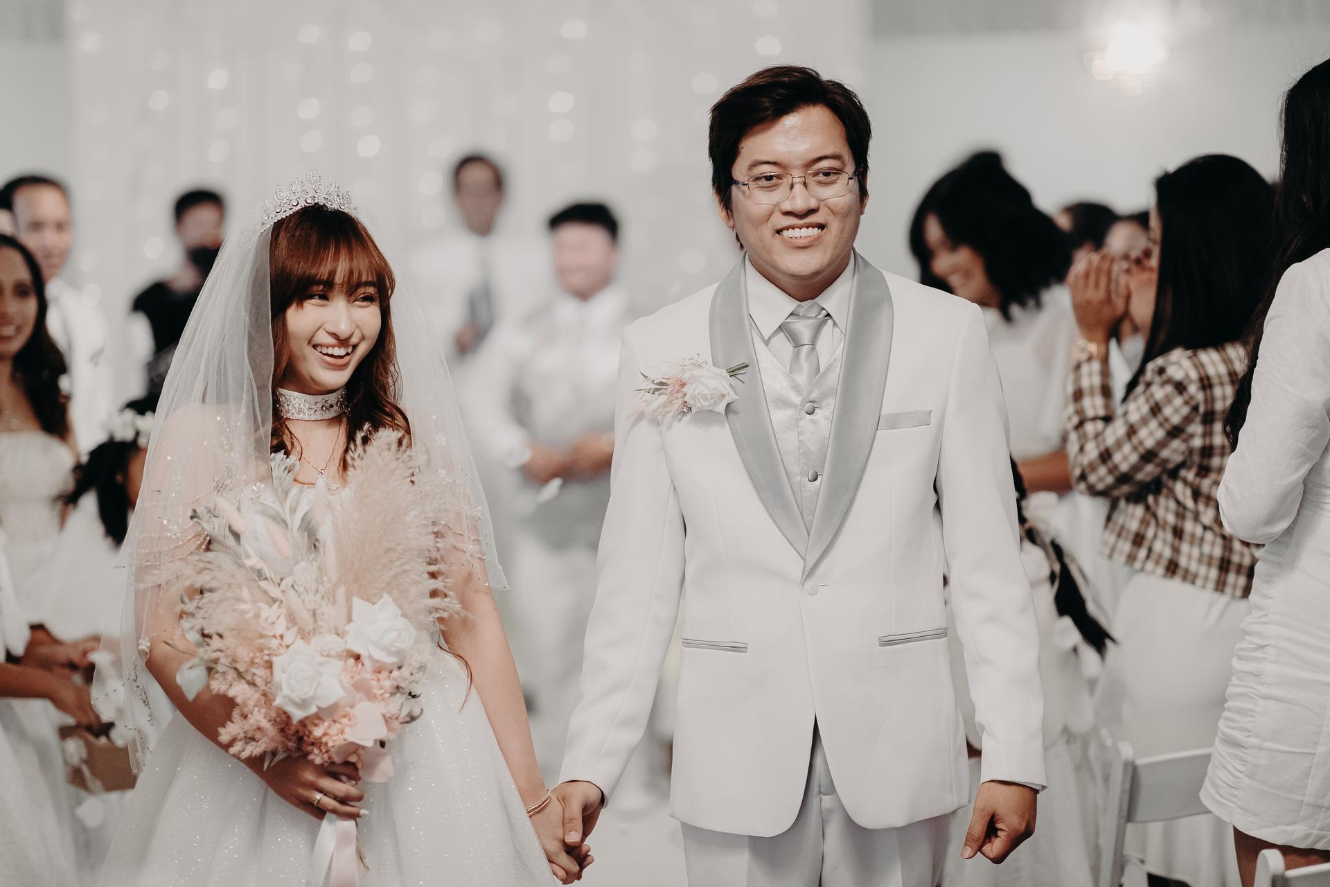 Bride Henny and groom Timothy walk down the aisle holding hands at their wedding ceremony at Royal on the Park, with guests standing and clapping on either side.