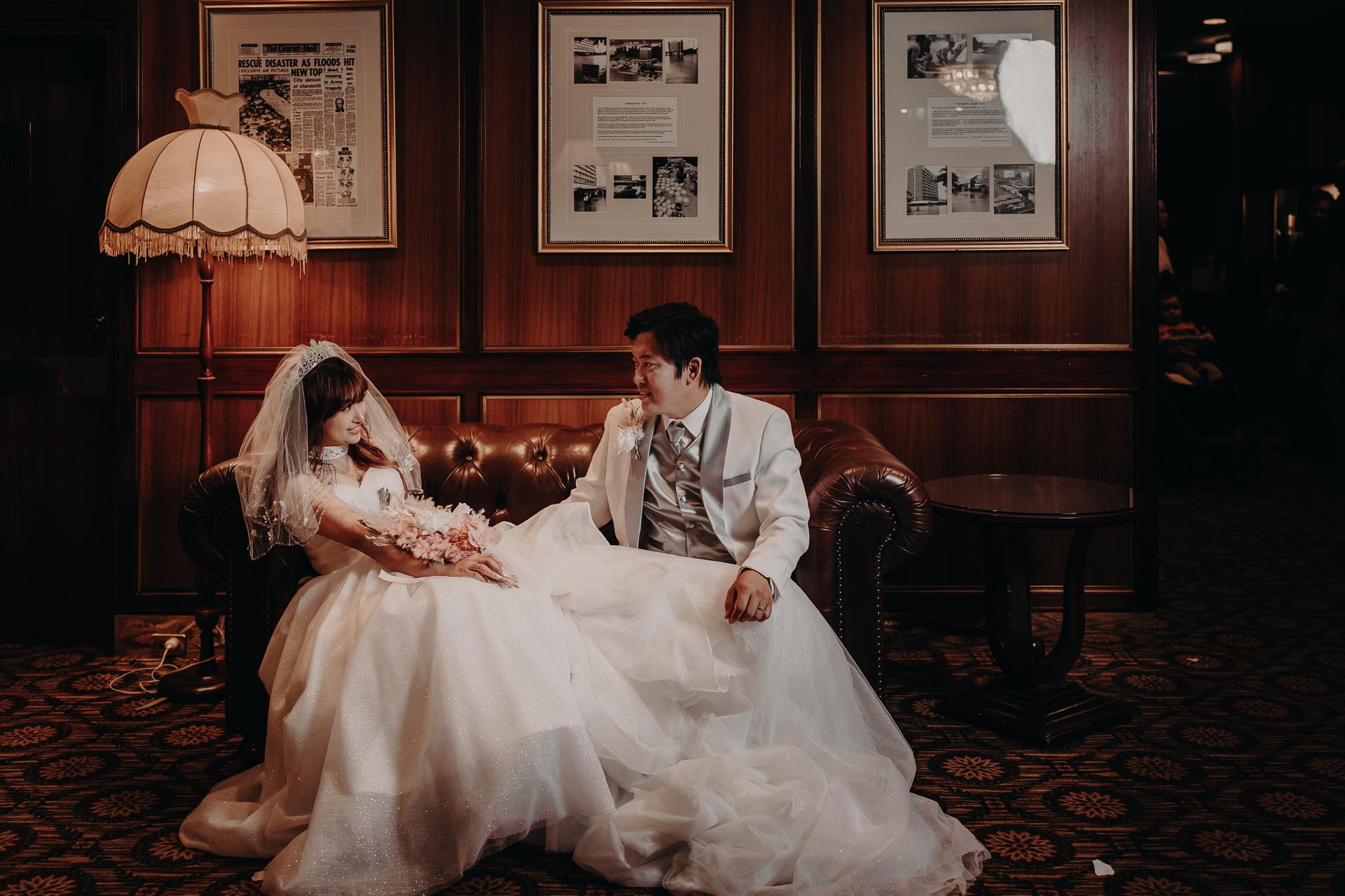 The bride Henny and groom Timothy sit together on a brown leather couch at Royal on the Park, with the bride holding a bouquet and the groom dressed in a white suit.
