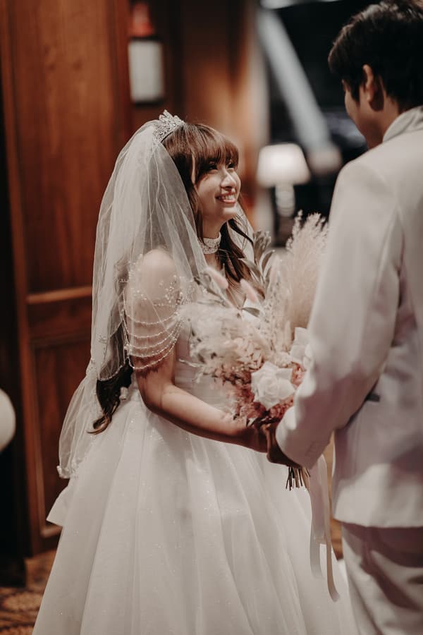The bride Henny in a white wedding gown and veil holding a bouquet smiles at the groom Timothy, who is dressed in a white suit, inside the Royal on the Park.
