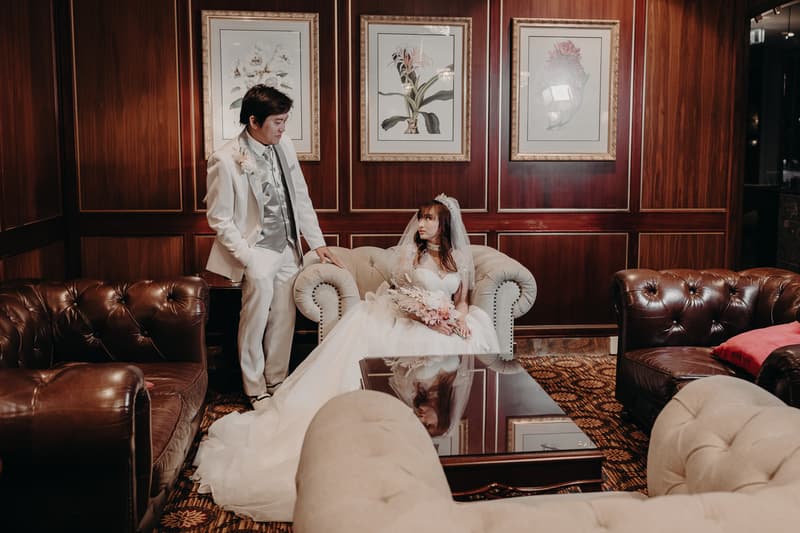 The bride Henny sits on a beige tufted sofa holding a bouquet, while the groom Timothy stands beside her in a white suit with a silver vest and tie, in a wood-paneled room at Royal on the Park.
