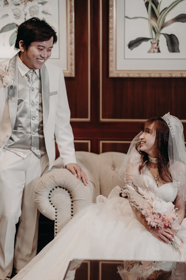 The groom Timothy in a white suit with a silver vest and tie stands smiling beside the bride Henny, who is seated on a beige tufted sofa wearing a white wedding gown and veil, holding a bouquet of flowers at Royal on the Park.