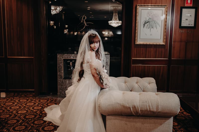 The bride Henny in a white wedding gown and veil poses indoors leaning on a beige tufted sofa at Royal on the Park.