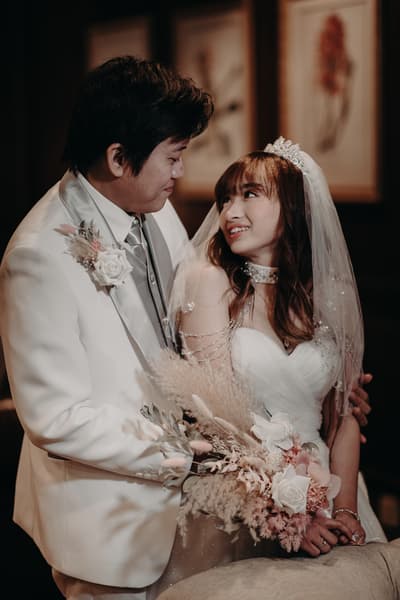 Timothy and Henny pose together holding a bouquet at Royal on the Park during their couple portraits session. Timothy wears a white suit with a boutonniere, and Henny wears a white wedding gown with a veil and tiara.