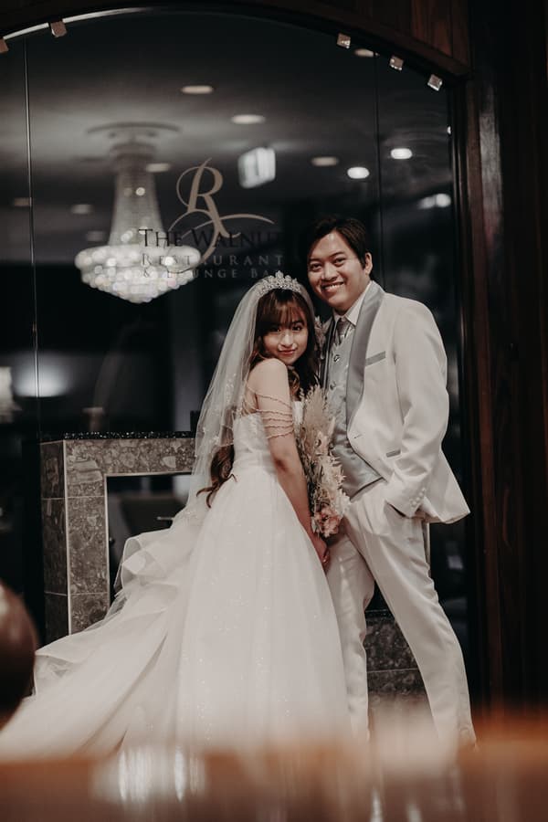 Bride Henny in a white wedding gown and veil holding a bouquet stands next to groom Timothy in a white suit with a silver vest and tie inside The Walnut Restaurant & Lounge Bar at Royal on the Park.