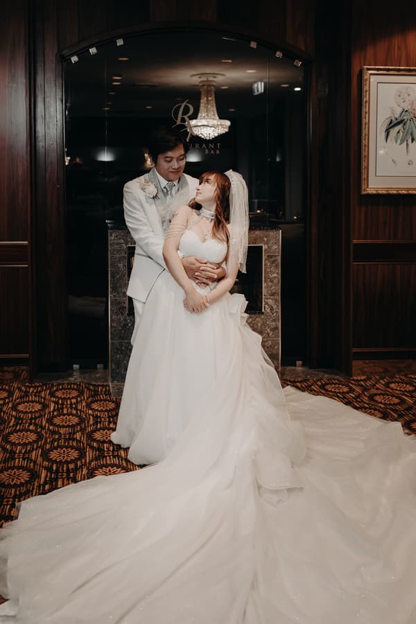 The groom Timothy embraces the bride Henny from behind as they pose for a portrait inside Royal on the Park, with a chandelier and dark wood paneling in the background.