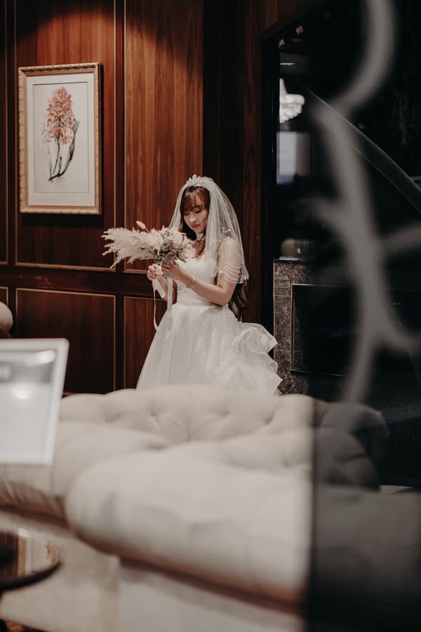 The bride Henny stands indoors at Royal on the Park wearing a white wedding gown and veil, holding a bouquet of dried flowers.