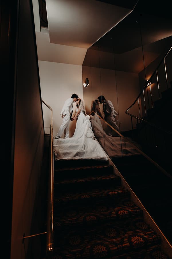 The bride and groom, Henny and Timothy, stand on a carpeted staircase at Royal on the Park, holding hands and leaning in for a kiss. The bride wears a long white gown with a train and veil, while the groom wears a white suit. Their reflection is visible in the mirrored wall beside them.