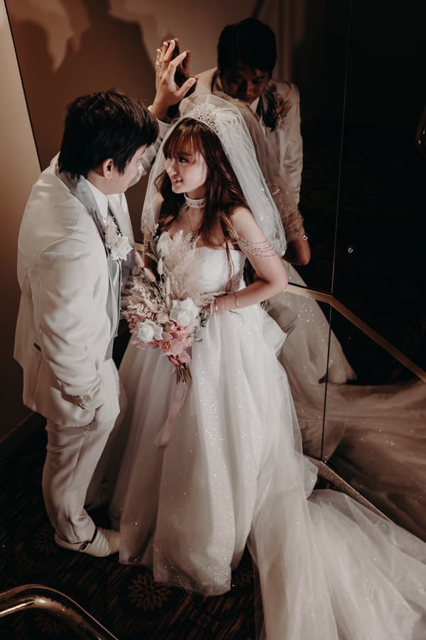 The bride Henny in a sparkling white gown and veil holds a bouquet while standing close to the groom Timothy, who is dressed in a white suit, near a mirrored wall at Royal on the Park.