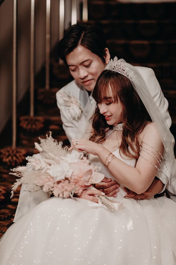 The bride Henny and groom Timothy sit closely together on a staircase at Royal on the Park, with Timothy embracing Henny from behind as she holds a bouquet of flowers.