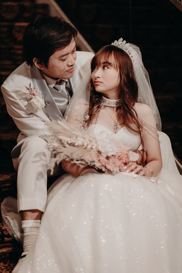 Timothy and Henny sit on carpeted stairs at Royal on the Park, with Timothy in a white suit and Henny in a white wedding gown and veil holding a bouquet.