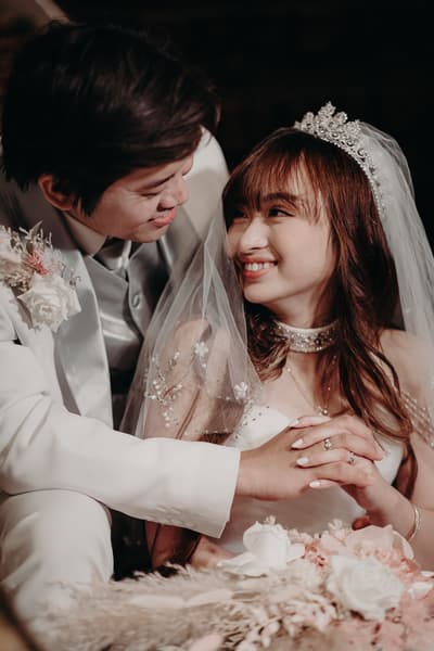 The groom Timothy in a white suit and the bride Henny in a white wedding gown with a tiara and veil smile at each other while holding hands, with a bouquet of flowers in the foreground. Photo and video services by She Said Yes.