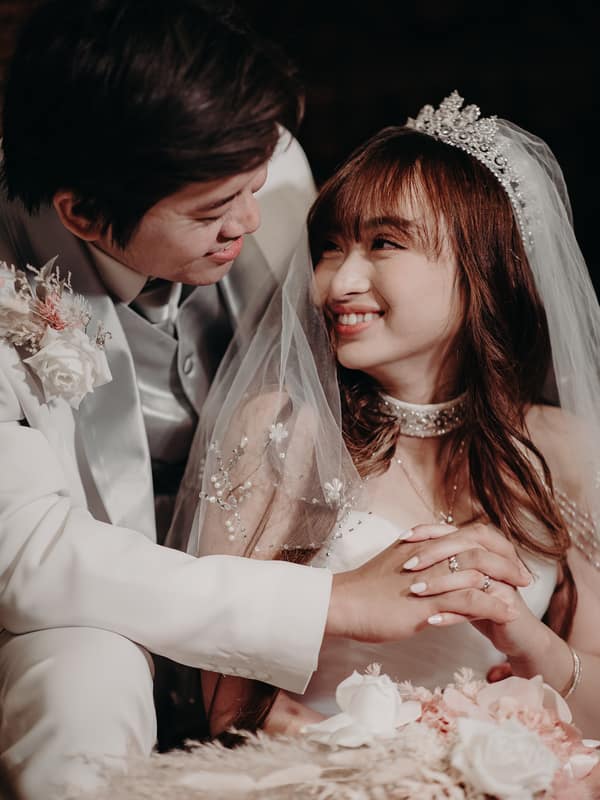 The groom Timothy in a white suit and the bride Henny in a white wedding gown with a tiara and veil smile at each other while holding hands, with a bouquet of flowers in the foreground at Royal on the Park.