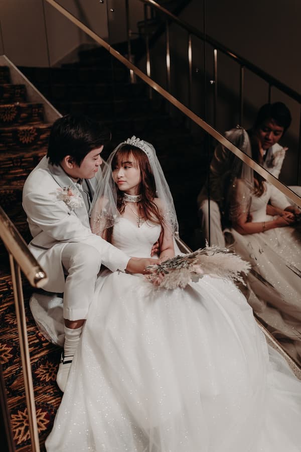 The bride Henny in a white wedding gown and veil sits on carpeted stairs next to the groom Timothy in a white suit at Royal on the Park, with their reflection visible in a glass panel beside them.