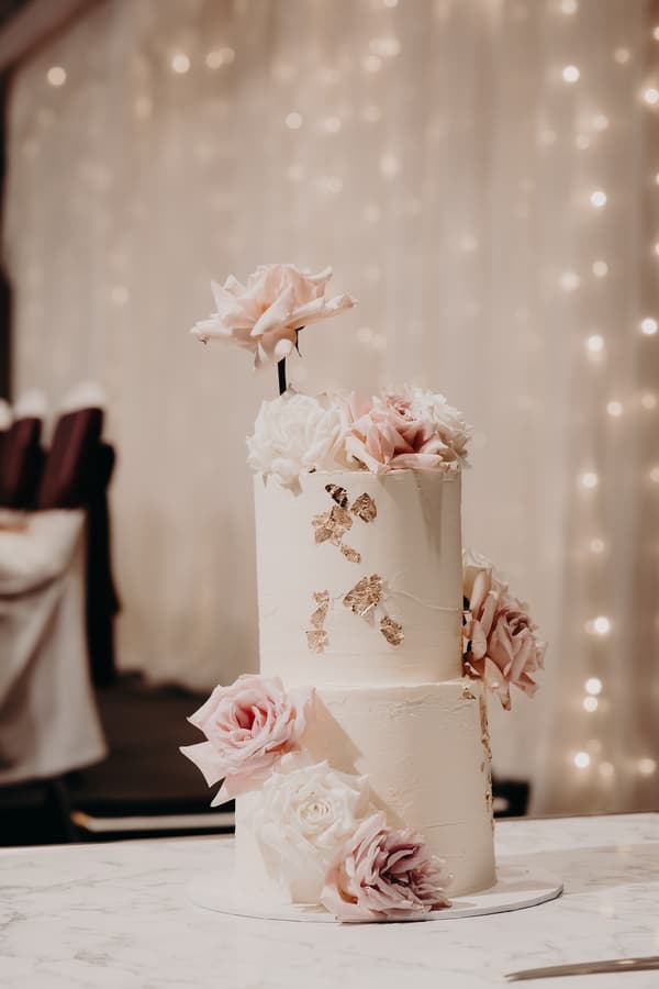 Two-tier white wedding cake decorated with pink and white roses and gold leaf accents on a marble table at Royal on the Park.
