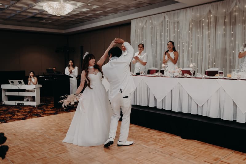 Bride Henny and groom Timothy dance on the parquet floor at the Royal on the Park reception while guests and bridal party members applaud from the head table and nearby.