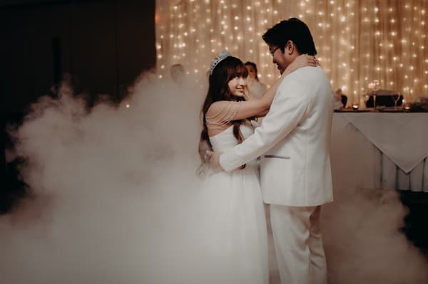 Bride Henny and groom Timothy share a dance surrounded by fog at the reception stage of Royal on the Park, with a backdrop of string lights and a decorated table.