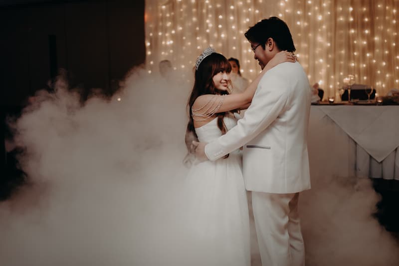 Bride Henny and groom Timothy share a dance surrounded by fog at the reception stage of Royal on the Park, with a backdrop of string lights and a decorated table.