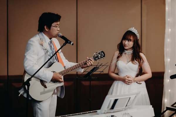 Timothy in a white suit plays guitar and sings into a microphone while Henny in a white wedding gown and tiara watches during the reception at Royal on the Park.