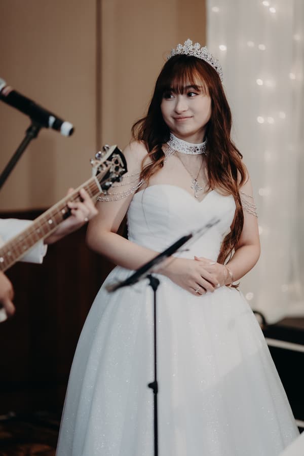The bride Henny stands near a microphone and a guitar player at the reception stage of Royal on the Park, wearing a white strapless gown with a tiara and jewelry.