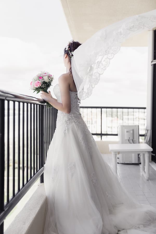 The bride stands alone on a balcony at Bilinga Beach Weddings, holding a bouquet of pink and white flowers, with her veil flowing behind her.