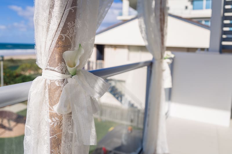 Close-up of white lace curtains tied with white ribbon and decorated with a white calla lily on a balcony railing, with a blurred view of buildings and the beach in the background. Sandstone point hotel wedding by She Said Yes.