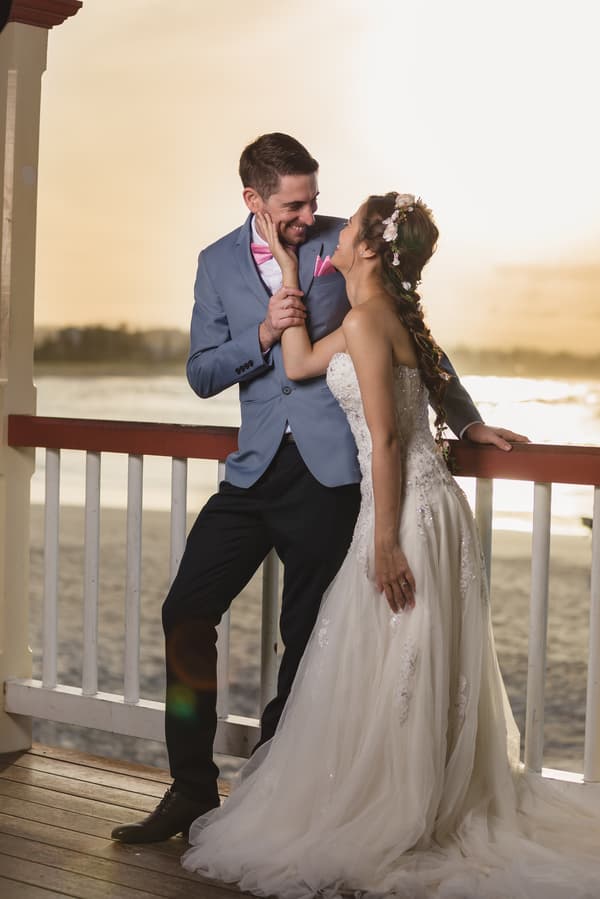 The bride Wing in a white strapless wedding gown with floral hair accessories and the groom Jason in a light blue suit jacket with a pink bow tie and pocket square pose together on a wooden deck railing overlooking the beach at Bilinga Beach Weddings during sunset.