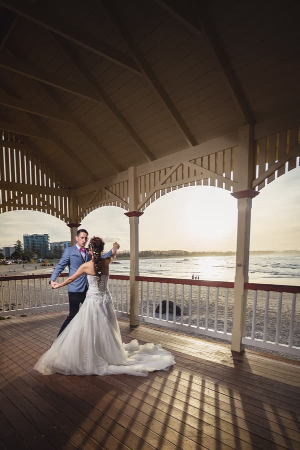 The bride and groom dance together under a wooden pavilion overlooking the beach at Bilinga Beach Weddings during sunset.