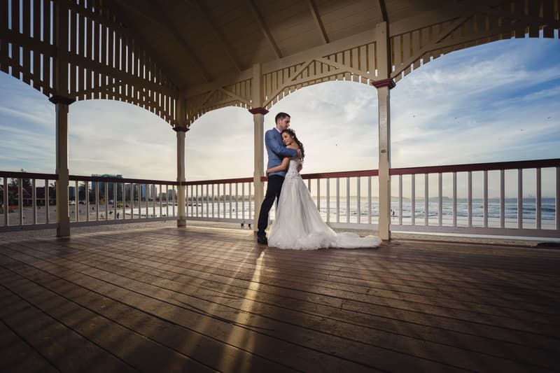 The bride and groom embrace under a wooden pavilion overlooking the beach at Bilinga Beach Weddings during a couple portrait session at sunset.