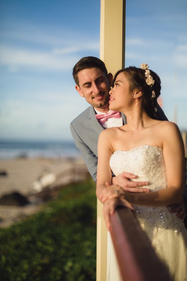 Bride Wing in a strapless white wedding gown with floral hair accessories and groom Jason in a light grey suit with a pink bow tie embrace near a wooden railing at Bilinga Beach Weddings with the beach and ocean in the background.