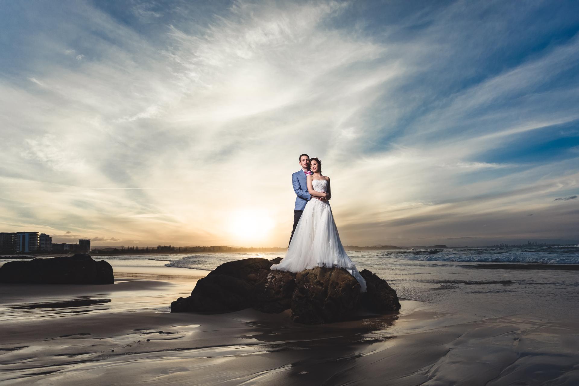 The bride Wing and groom Jason stand together on a large rock at Bilinga Beach Weddings during sunset, with the ocean and sky in the background.