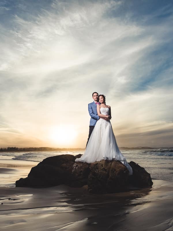 The bride Wing and groom Jason stand together on a large rock at Bilinga Beach Weddings during sunset, with the ocean and sky in the background.