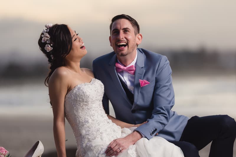 The bride Wing and groom Jason sit closely together on the beach at Bilinga Beach Weddings, both laughing and dressed in wedding attire; Wing wears a strapless white lace gown with floral hair accessories, and Jason wears a blue suit with a pink bow tie and pocket square.